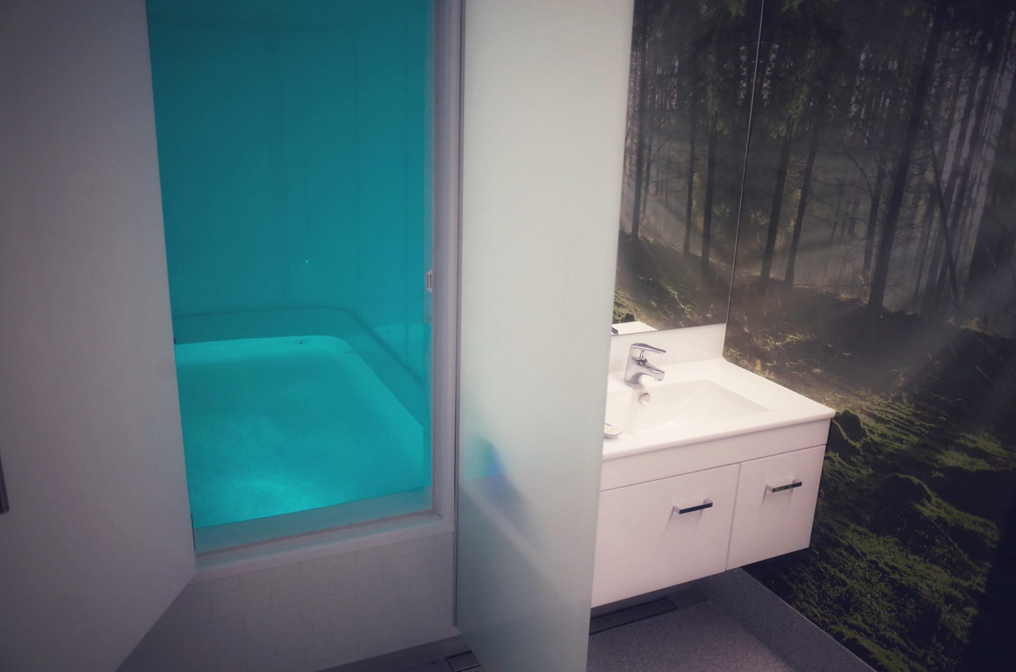 The Titirangi float room — a private float tank lit in calm teal, beside a forest-scene wall